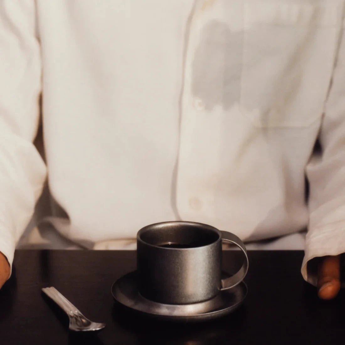 Kaffekopp med fat i borstat rostfritt stål och en sked liggandes bredvid.