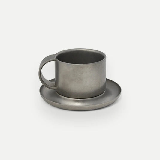 Kaffekopp i borstat rostfritt stål med fat mot vit bakgrund