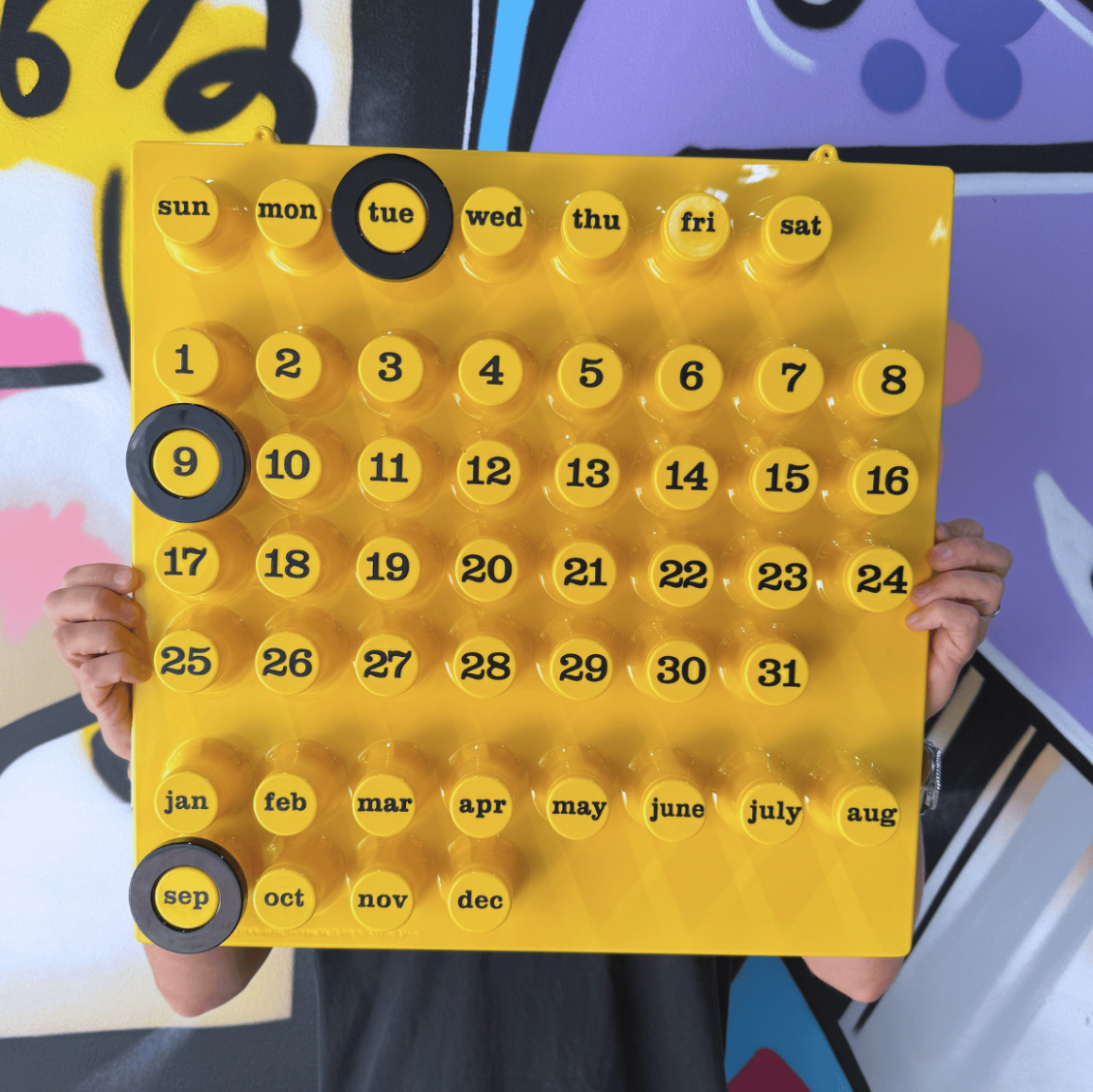 Gul väggkalender med 3 svarta ringar. En man som håller ståendes mot en graffitivägg