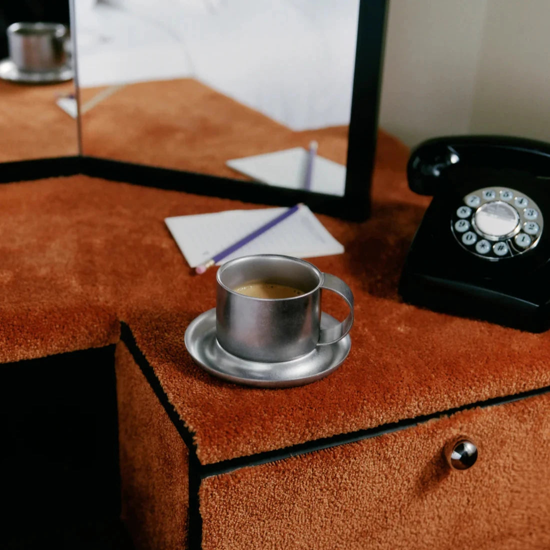 Kaffekopp med fat i borstat rostfritt stål från Service Projects. På bilden står den på en möbel inklädd byrå med en gammaldags svart telefon på. I bakgrunden en spegel, ett block och en penna. Bilden är i lite retrostil