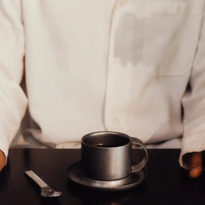 Kaffekopp med fat i borstat rostfritt stål och en sked liggandes bredvid.