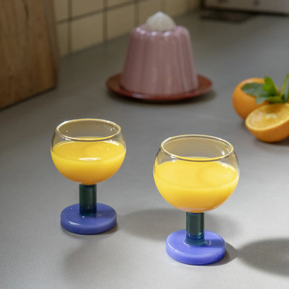 2 glas på fot från &Klevering med apelsinjuice i. Foten på glasen är i grönt och blått. 