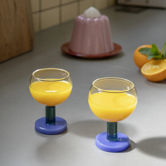 2 glas på fot från &Klevering med apelsinjuice i. Foten på glasen är i grönt och blått. 