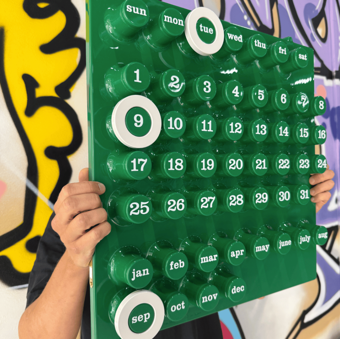 Grön väggkalender med vita ringar mot grafittibakgrund. En kille som håller i kalendern