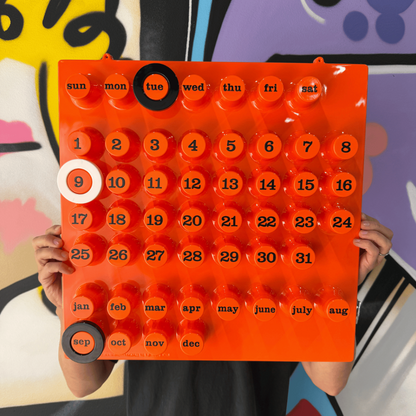 Italiensk väggkalender i orange med två svarta och en vit ring. En man som håller ståendes mot en graffitivägg.